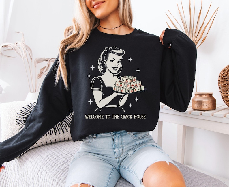 Op de afbeelding: Zwart sweatshirt met lange mouwen met een retro-illustratie van een vrouw die een dienblad met speltegels vasthoudt. De tekst "WELCOME TO THE CRACK HOUSE" staat eronder. Het sweatshirt wordt door een persoon gedragen.