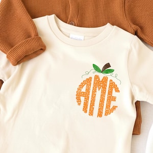 May include: Cream-colored t-shirt with a pumpkin design. The pumpkin is orange with the letters "AME" inside. A brown ribbed long-sleeve shirt is visible in the background.