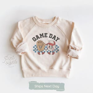 May include: Cream-colored sweatshirt with raglan sleeves. The front features a graphic of a baseball glove and baseball character with the words "GAME DAY" above them. The design is set against a checkered background. The sweatshirt has a crew neck.