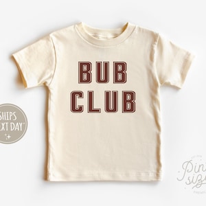 May include: A cream colored short-sleeved t-shirt with the text "BUB CLUB" printed in brown.