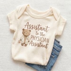 May include: Cream-colored baby onesie with the text "Assistant to the Physician Assistant" in brown script. A cartoon bear wearing a doctor's coat and holding a medical bag is printed on the front. Blue denim jeans are visible in the bottom right corner.