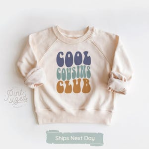 May include: Cream-colored sweatshirt with raglan sleeves. The front features the text "COOL COUSINS CLUB" in blue, teal, and brown retro font. The sweatshirt is a casual style, suitable for children's wear.