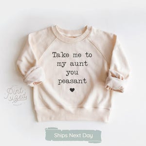 May include: Cream-colored sweatshirt with raglan sleeves. The front features the text "Take me to my aunt you peasant" in black, with a small black heart below. The sweatshirt has a crew neck and ribbed cuffs.