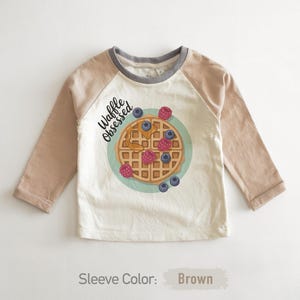 Peut inclure: Un t-shirt blanc &agrave; manches longues avec des manches marron et un col gris. Il pr&eacute;sente un dessin de gaufre avec des baies et le texte "Waffle Obsessed". Un design amusant pour enfants.