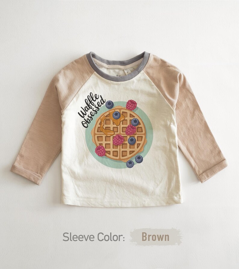 Peut inclure: Un t-shirt blanc &agrave; manches longues avec des manches marron et un col gris. Il pr&eacute;sente un dessin de gaufre avec des baies et le texte "Waffle Obsessed". Un design amusant pour enfants.