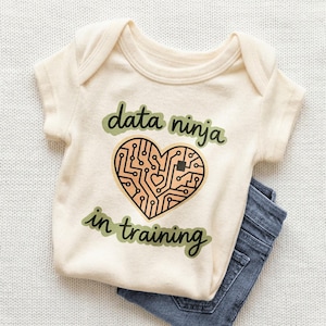 May include: Cream-colored baby onesie with the text "data ninja in training" in green. A heart-shaped circuit board design is in the center. Blue denim jeans are visible on the side.