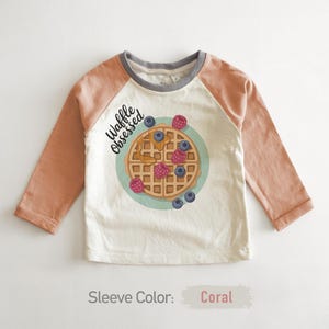 Peut inclure: Un t-shirt &agrave; manches longues blanc cass&eacute; avec des manches corail et un col gris. Le t-shirt pr&eacute;sente un motif de gaufre avec des baies et le texte "Waffle Obsessed". La couleur des manches est corail.