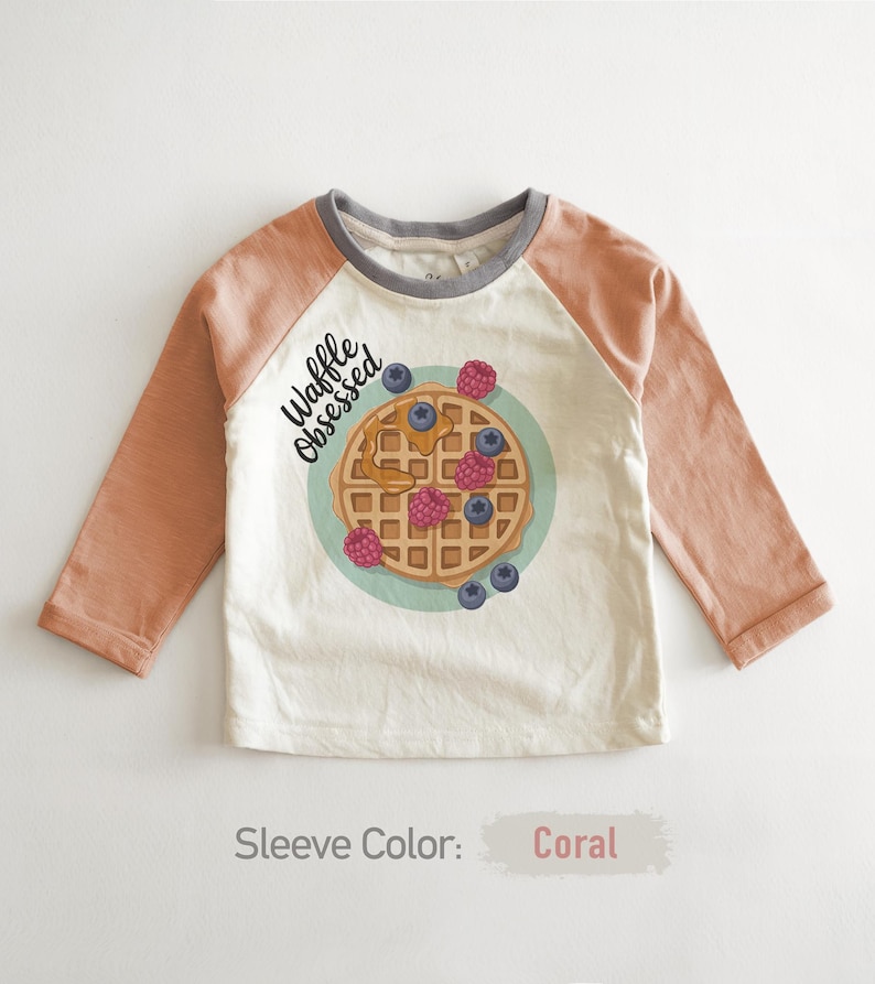 Peut inclure: Un t-shirt &agrave; manches longues blanc cass&eacute; avec des manches corail et un col gris. Le t-shirt pr&eacute;sente un motif de gaufre avec des baies et le texte "Waffle Obsessed". La couleur des manches est corail.