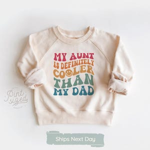 May include: Cream-colored sweatshirt with raglan sleeves. The front features the text "MY AUNT IS DEFINITELY COOLER THAN MY DAD" in retro-style lettering. The text is in shades of red, orange, yellow, teal, and blue. The sleeves are cuffed.