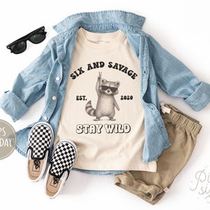 May include: A cream-colored t-shirt with the text "SIX AND SAVAGE EST. 2020 STAY WILD" and a raccoon graphic wearing sunglasses. The shirt is paired with khaki shorts, a denim shirt, and black and white checkered slip-on shoes. Black sunglasses are also included.
