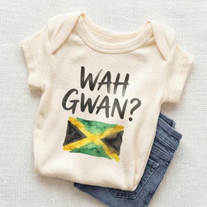 May include: A cream-colored baby onesie with the text "WAH GWAN?" in black lettering. Below the text is a watercolor-style Jamaican flag. Blue denim jeans are partially visible.