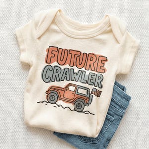 May include: Cream-colored baby onesie with the text "FUTURE CRAWLER" in orange and gray. A cartoon jeep with an American flag is printed below the text. Blue denim jeans are folded beneath the onesie.