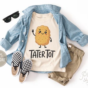 May include: A cream-colored t-shirt with a cartoon tater tot graphic and the text "TATER TOT". The shirt is paired with khaki shorts, a light blue denim shirt, and black and white checkered slip-on shoes. Black sunglasses are also included.