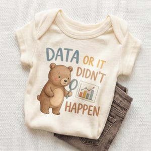 May include: Cream-colored baby onesie with the text "DATA OR IT DIDN'T HAPPEN" and a cartoon bear holding a magnifying glass. The bear is brown with a light tan belly. Brown pants are folded beneath the onesie.
