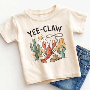 May include: Cream-colored t-shirt with a desert-themed graphic featuring a red lobster, cacti, a cowboy hat, and the text "YEE-CLAW". The design is set against a backdrop of a sun and desert landscape. Blue jeans are folded next to the shirt.