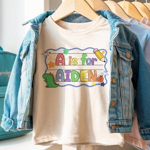 May include: A cream-colored t-shirt with the text "A is for AIDEN" in colorful letters, a dinosaur, a pencil, and an apple. The shirt is paired with a light blue denim jacket and is hanging on a wooden hanger.