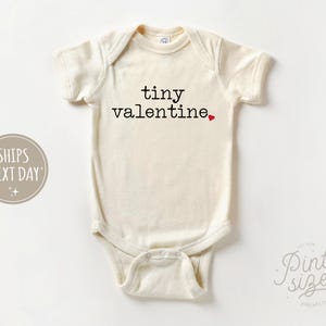 May include: A cream-colored baby onesie with the text "tiny valentine" in black lettering and a small red heart.