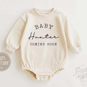 May include: Cream-colored baby onesie with long sleeves. The text "BABY Hunter COMING SOON" is printed in black script across the front. The onesie has a round neckline and snap closures at the bottom. A small logo is visible in the lower right corner.