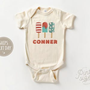 May include: A white baby onesie with a red, white, and blue popsicle graphic and the name "CONNER" printed in red.