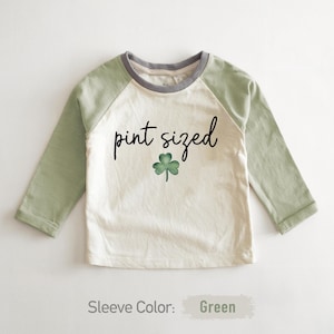May include: A white long-sleeve shirt with green sleeves. The shirt has a gray crew neck and a shamrock graphic with the text "pint sized".