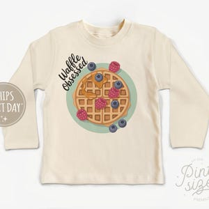 Peut inclure: T-shirt cr&egrave;me &agrave; manches longues avec un motif de gaufre, des myrtilles et des framboises. Le texte "Waffle Obsessed" est au-dessus du motif de la gaufre. Le t-shirt a un col rond et des manches longues.