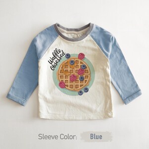 Peut inclure: T-shirt &agrave; manches longues avec des manches bleu clair et un corps cr&egrave;me. Il pr&eacute;sente un motif de gaufre avec des baies et le texte "Waffle Obsessed". Le col est gris.