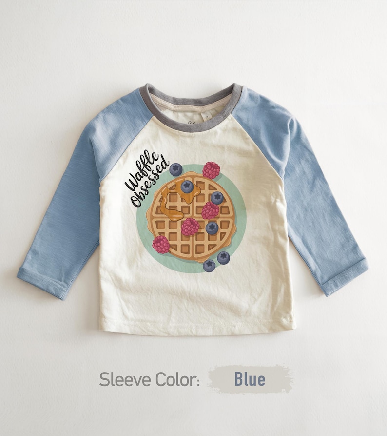 Peut inclure: T-shirt &agrave; manches longues avec des manches bleu clair et un corps cr&egrave;me. Il pr&eacute;sente un motif de gaufre avec des baies et le texte "Waffle Obsessed". Le col est gris.
