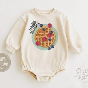 Peut inclure: Barboteuse b&eacute;b&eacute; cr&egrave;me avec un motif de gaufre orn&eacute;e de myrtilles et de framboises sur une assiette turquoise. L'inscription "Waffle Obsessed" est au-dessus. La barboteuse a des manches longues et des boutons-pression.