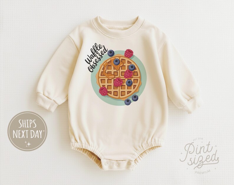 Peut inclure: Barboteuse b&eacute;b&eacute; cr&egrave;me avec un motif de gaufre orn&eacute;e de myrtilles et de framboises sur une assiette turquoise. L'inscription "Waffle Obsessed" est au-dessus. La barboteuse a des manches longues et des boutons-pression.