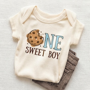 May include: Cream-colored baby onesie with a brown cookie graphic and the words "ONE SWEET BOY" in brown and blue. Brown denim pants are folded beneath the onesie. The onesie has short sleeves and a lap shoulder neckline.