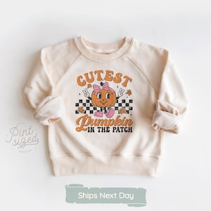May include: A cream colored sweatshirt with a graphic of a smiling pumpkin wearing a pink bow and the text "Cutest Pumpkin in the Patch".