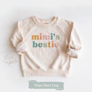 May include: Cream-colored sweatshirt with raglan sleeves. The front features the text "mimi's bestie" in colorful letters. The sweatshirt has a crew neck and rolled cuffs. The image also includes the text "Pint Sized Premium" and "Ships Next Day".