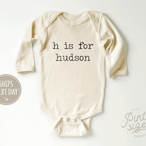 May include: A white long-sleeved baby bodysuit with the text "h is for hudson" printed in black.