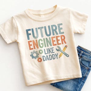 May include: Cream-colored short-sleeved t-shirt with the text "FUTURE ENGINEER LIKE DADDY" in blue, orange, and green. The design includes illustrations of gears, a wrench, and a pencil. Blue jeans are folded next to the shirt.