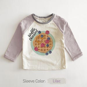Peut inclure: Un t-shirt blanc cass&eacute; &agrave; manches longues avec des manches lilas et un col gris. Il pr&eacute;sente un dessin de gaufre avec des baies et le texte "Waffle Obsessed".