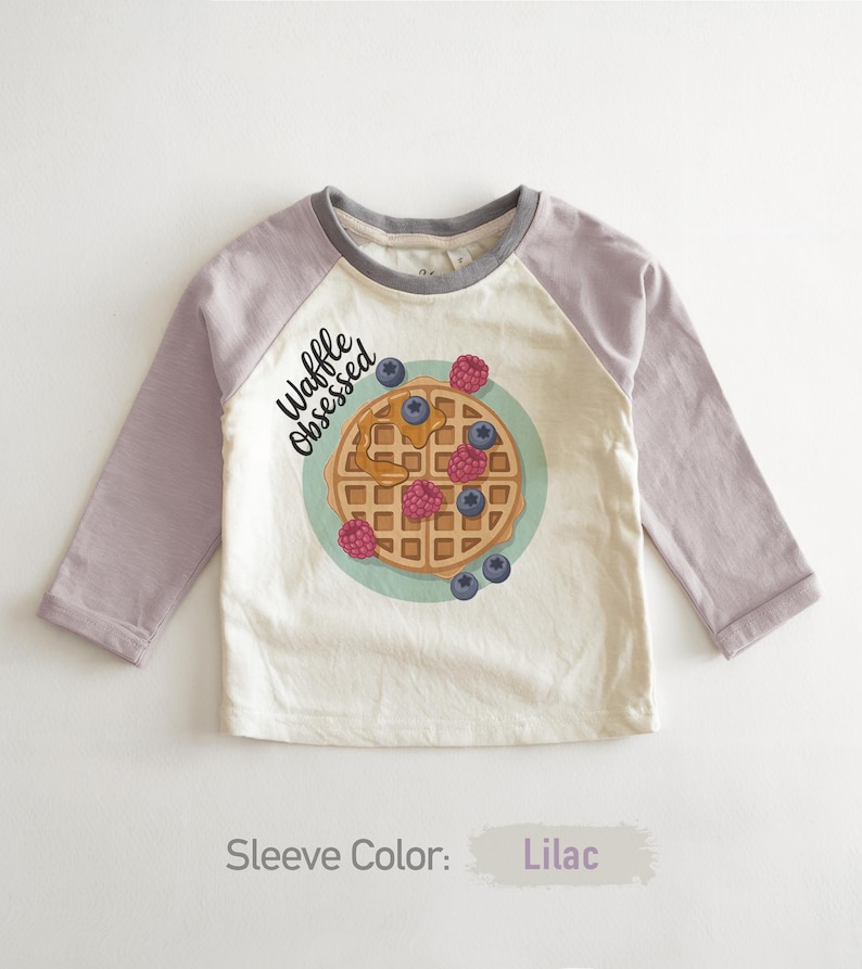 Peut inclure: Un t-shirt blanc cass&eacute; &agrave; manches longues avec des manches lilas et un col gris. Il pr&eacute;sente un dessin de gaufre avec des baies et le texte "Waffle Obsessed".