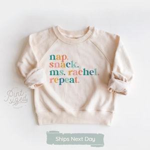 May include: Cream-colored sweatshirt with the text "nap. snack. ms. rachel. repeat." in colorful letters. The sweatshirt has long sleeves and a crew neck. The brand logo "Pint Sized Premium" is visible on the left side.