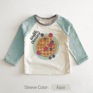 Peut inclure: Un t-shirt de baseball &agrave; manches longues avec des manches bleu-vert et un corps cr&egrave;me. Le t-shirt pr&eacute;sente un dessin de gaufre avec des baies et le texte "Waffle Obsessed". Le col est gris.