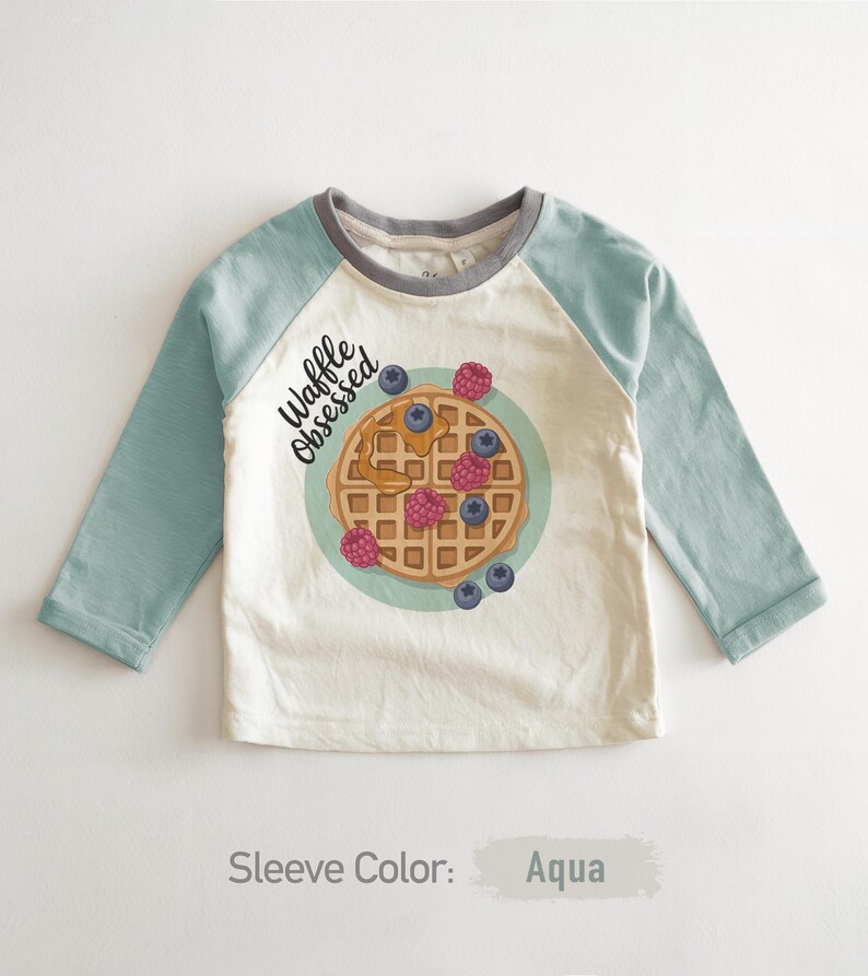 Peut inclure: Un t-shirt de baseball &agrave; manches longues avec des manches bleu-vert et un corps cr&egrave;me. Le t-shirt pr&eacute;sente un dessin de gaufre avec des baies et le texte "Waffle Obsessed". Le col est gris.