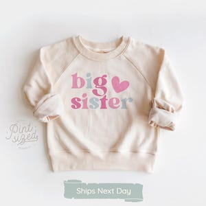 May include: Cream-colored sweatshirt with raglan sleeves. The front features the words "big sister" in pink, blue, and a pink heart. The sleeves are cuffed. The text "Ships Next Day" is at the bottom.