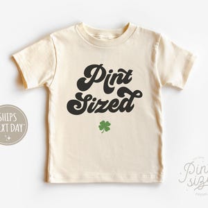 May include: A cream colored short-sleeved t-shirt with the text "Pint Sized" in black retro font. A green shamrock is below the text.