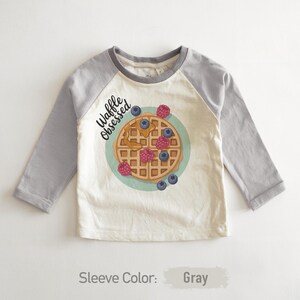 Peut inclure: Un t-shirt raglan &agrave; manches longues avec un corps cr&egrave;me et des manches grises. Le t-shirt pr&eacute;sente un motif de gaufre avec des myrtilles et des framboises, et les mots "Waffle Obsessed".