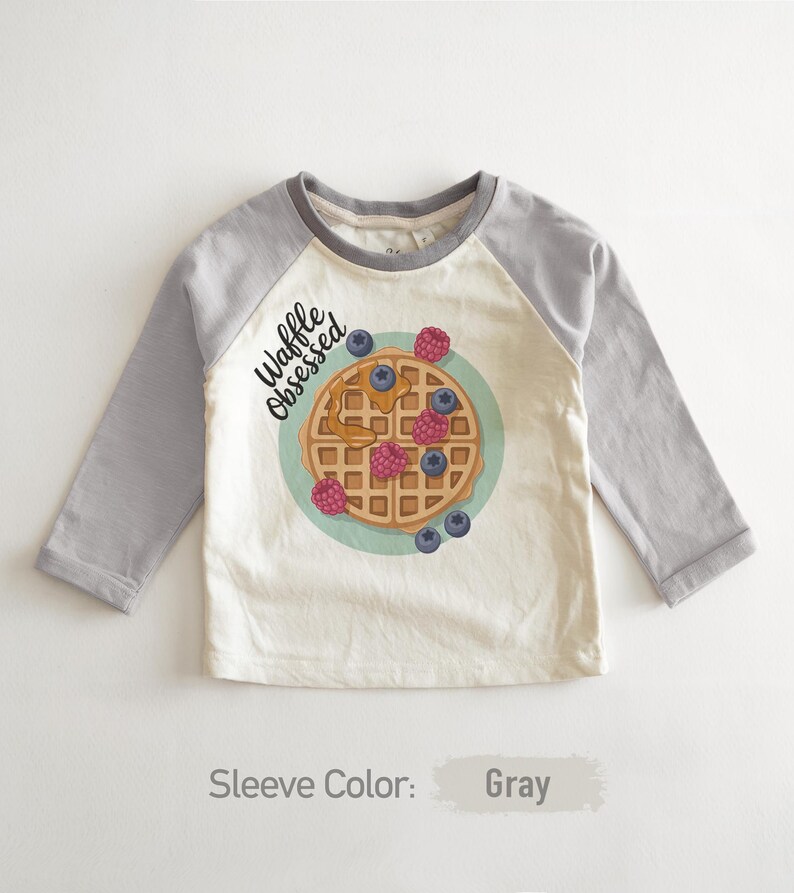 Peut inclure: Un t-shirt raglan &agrave; manches longues avec un corps cr&egrave;me et des manches grises. Le t-shirt pr&eacute;sente un motif de gaufre avec des myrtilles et des framboises, et les mots "Waffle Obsessed".