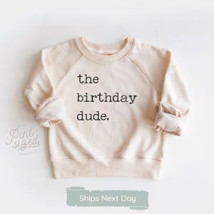 May include: Cream-colored sweatshirt with the text "the birthday dude." in black. The raglan sleeves and cuffs are rolled up. The sweatshirt is on a white background.