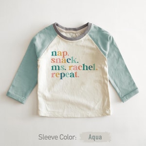 May include: A white long-sleeved t-shirt with aqua blue sleeves. The shirt has a colorful graphic that says "nap. snack. ms. rachel. repeat."