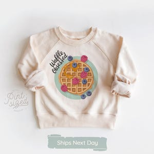 Peut inclure: Sweat-shirt cr&egrave;me avec un motif de gaufre. La gaufre est garnie de baies et de sirop, entour&eacute;e d'un cercle bleu clair. Le texte "Waffle Obsessed" est au-dessus du motif. Les manches sont retrouss&eacute;es.