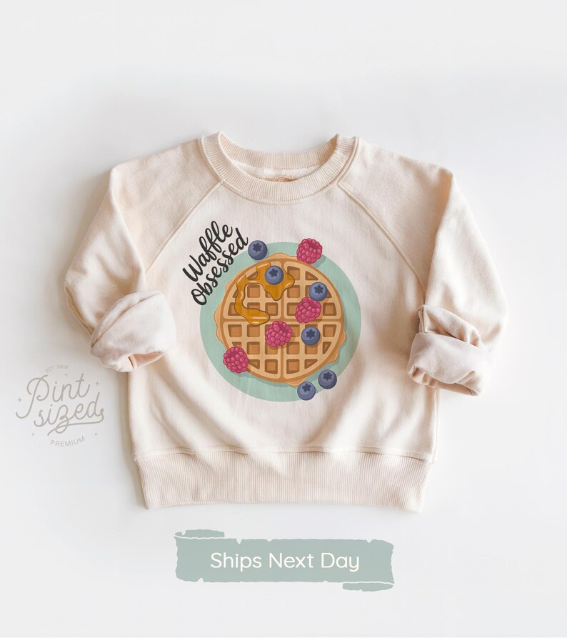 Peut inclure: Sweat-shirt cr&egrave;me avec un motif de gaufre. La gaufre est garnie de baies et de sirop, entour&eacute;e d'un cercle bleu clair. Le texte "Waffle Obsessed" est au-dessus du motif. Les manches sont retrouss&eacute;es.