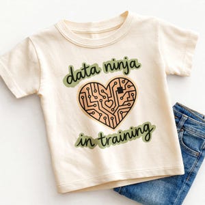 May include: Cream-colored t-shirt with the text "data ninja in training" in green script. A heart-shaped circuit board design is in the center. Blue jeans are folded next to the shirt.