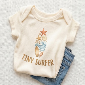 May include: Cream-colored baby onesie with a watercolor design of a surfboard, waves, starfish, and seashells. The words "TINY SURFER" are printed in brown script. Blue denim jeans are folded next to the onesie.