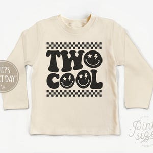 May include: A cream colored long-sleeved shirt with a black graphic that says "TWO COOL" with two smiley faces with lightning bolts for eyes. The graphic is framed by a black and white checkered pattern.